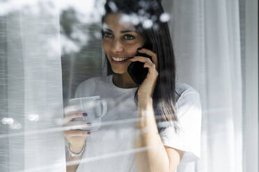 Lächelnde junge Frau am Mobiltelefon hinter einer Fensterscheibe - GIOF06948