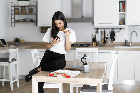 Young woman with cell phone eating pizza in kitchen at home stock photo