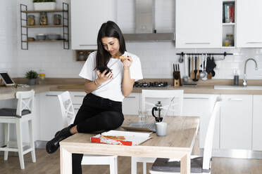 Junge Frau mit Mobiltelefon isst Pizza in der Küche zu Hause - GIOF06949