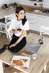 Junge Frau mit Mobiltelefon isst Pizza in der Küche zu Hause - GIOF06950