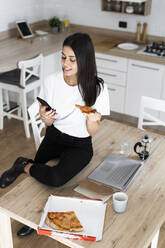 Junge Frau mit Mobiltelefon isst Pizza in der Küche zu Hause - GIOF06951
