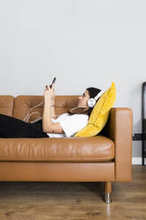 Junge Frau auf der Couch zu Hause mit Smartphone und Kopfhörern liegend - GIOF06956