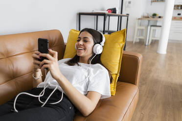 Young woman lying on the couch at home with smartphone and headphones - GIOF06957