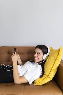 Junge Frau auf der Couch zu Hause mit Smartphone und Kopfhörern liegend - GIOF06958