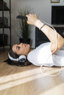 Junge Frau liegt mit Smartphone und Kopfhörern auf dem Boden - GIOF06961
