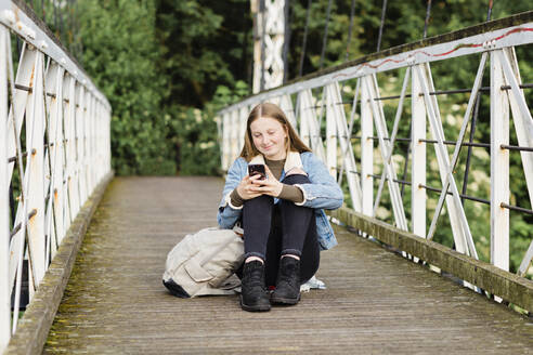 Teenager-Mädchen sitzt auf einer Brücke und benutzt ein Smartphone - NMS00365