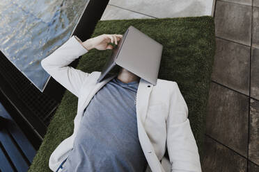 Junger Geschäftsmann entspannt sich auf Kunstrasen und bedeckt sein Gesicht mit seinem Laptop - KMKF01025