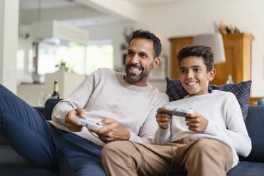 Happy father and son playing video game on couch in living room - DIGF07723