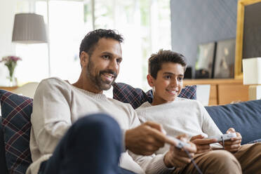 Happy father and son playing video game on couch in living room - DIGF07726