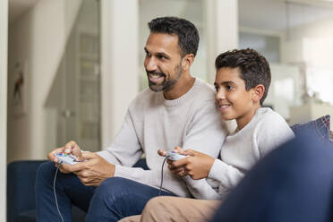 Happy father and son playing video game on couch in living room - DIGF07728