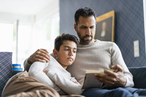Vater liegt mit Sohn auf der Couch im Wohnzimmer und liest ein Buch, lizenzfreies Stockfoto