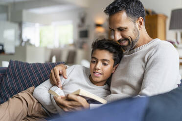 Father lying with son on couch in living room reading book - DIGF07730