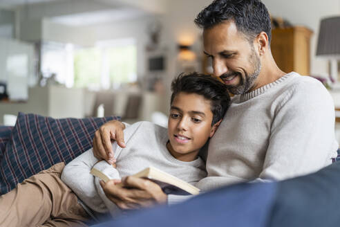 Vater liegt mit Sohn auf der Couch im Wohnzimmer und liest ein Buch - DIGF07730