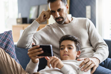 Father and son using smartphone on couch in living room - DIGF07731
