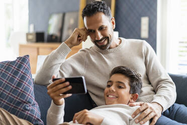 Smiling father and son using smartphone on couch in living room - DIGF07732