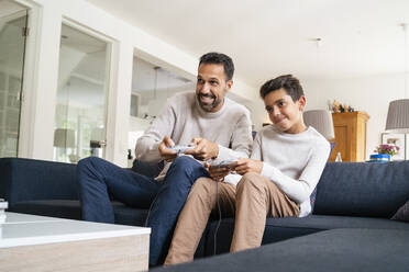 Happy father and son playing video game on couch in living room - DIGF07758