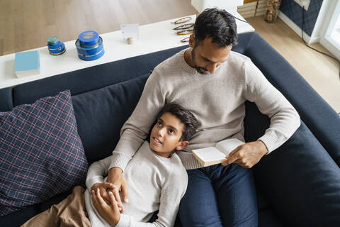 Vater liegt mit Sohn auf der Couch im Wohnzimmer und liest ein Buch, lizenzfreies Stockfoto