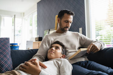 Father lying with son on couch in living room reading book - DIGF07760