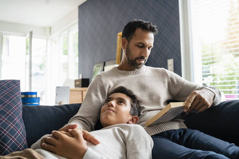 Vater liegt mit Sohn auf der Couch im Wohnzimmer und liest ein Buch - DIGF07760