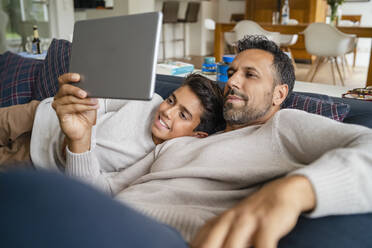 Happy father and son using tablet on couch in living room - DIGF07761