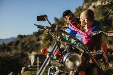 Happy gay couple with gay pride flag on a motorbike - LJF00490