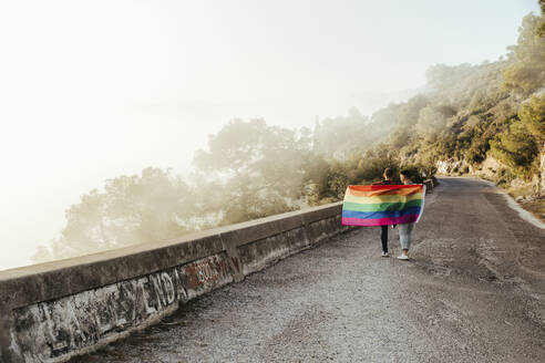Gay couple with gay pride flag on a road in the mountains - LJF00497