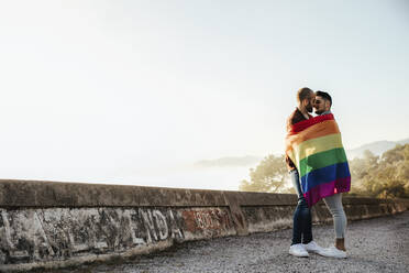 Homosexuelles Paar, eingewickelt in eine Gay Pride Flagge, küsst sich auf einer Straße in den Bergen - LJF00499