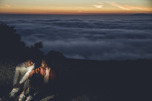 Homosexuelles Paar sitzt in den Bergen über den Wolken bei Sonnenuntergang - LJF00516