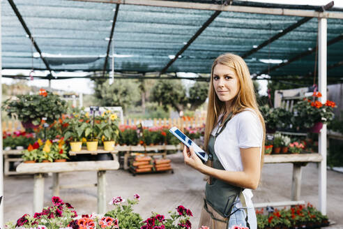 Portrait of a smiling female worker in a garden center holding a tablet - JRFF03514