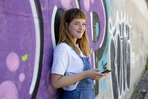 Lächelnde junge Frau mit Smartphone vor einer lila Graffiti-Wand - FLLF00250