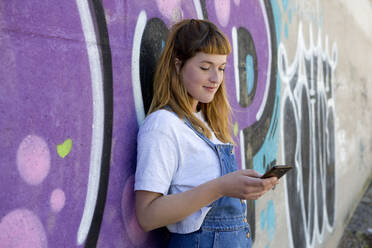 Junge Frau mit Smartphone vor einer lila Graffiti-Wand - FLLF00251