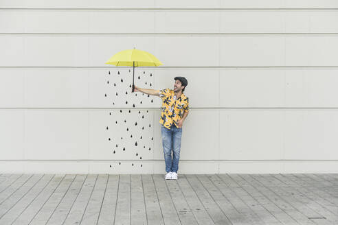 Digitales Komposit eines jungen Mannes, der einen Regenschirm an eine Wand mit Regentropfen hält - UUF18362