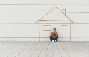 Digital composite of young man sitting at a wall with a house - UUF18365