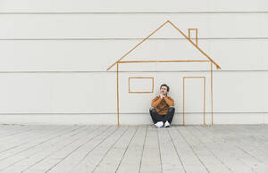 Digitales Komposit eines jungen Mannes, der an einer Mauer mit einem Haus sitzt - UUF18365