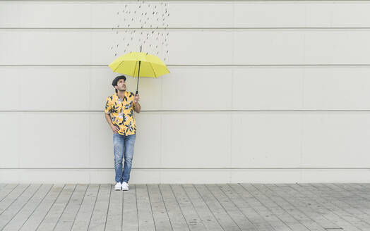 Digitales Komposit eines jungen Mannes, der einen Regenschirm an eine Wand mit Regentropfen hält - UUF18369