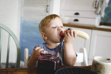 Cute little boy eating banana with blueberry jam in the kitchen - EYAF00333