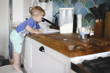 Kleiner Junge steht auf Zehenspitzen auf einem Stuhl in der Küche und wäscht Geschirr - EYAF00337