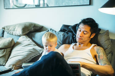 Father and son reading on sofa in living room - BLEF12849