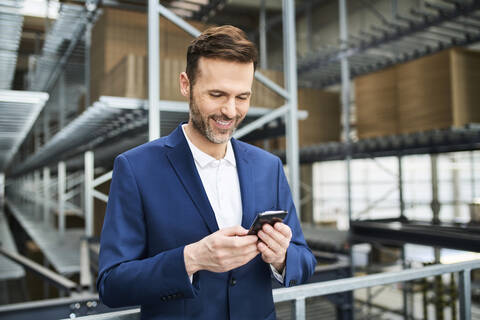 Lächelnder Geschäftsmann benutzt Handy in einer Fabrik, lizenzfreies Stockfoto