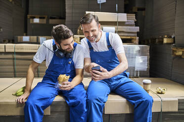 Zwei glückliche Arbeiter benutzen ein Handy während der Mittagspause in einer Fabrik - BSZF01218