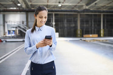 Lächelnde Geschäftsfrau mit Handy in einer Fabrik - BSZF01223