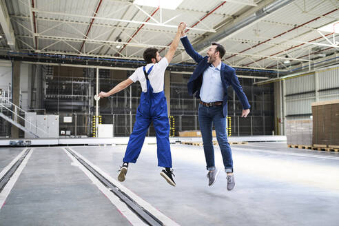 Excited businessman and worker jumping and high fiving in a factory - BSZF01249