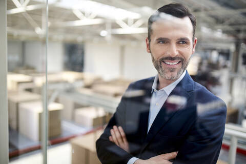 Porträt eines lächelnden Geschäftsmannes hinter einer Glasscheibe in einer Fabrik, lizenzfreies Stockfoto