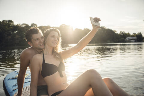 Glückliches Paar sitzt auf einem Paddelbrett auf einem See und macht Smartphone-Selfies, lizenzfreies Stockfoto