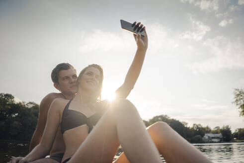 Glückliches Paar sitzt auf einem Paddelbrett auf einem See und macht Smartphone-Selfies - GUSF02314