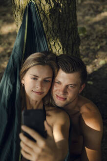 Ein Paar entspannt sich in einer Hängematte am See und macht Smartphone-Selfies - GUSF02332