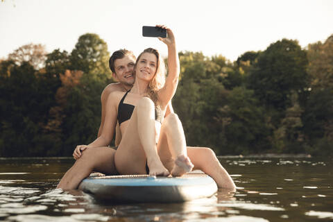 Glückliches Paar sitzt auf einem Paddelbrett auf einem See und macht Smartphone-Selfies, lizenzfreies Stockfoto