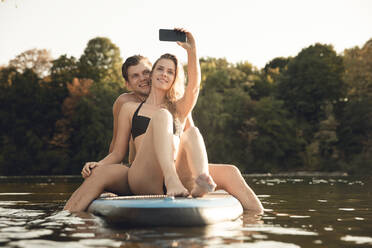 Glückliches Paar sitzt auf einem Paddelbrett auf einem See und macht Smartphone-Selfies - GUSF02351