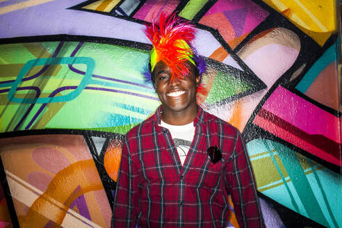 Black man in colorful wig smiling near graffiti wall - BLEF13131