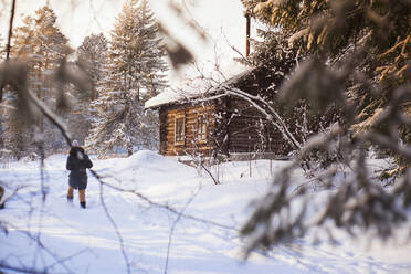 Kaukasische Frau geht in der Nähe einer Blockhütte im verschneiten Wald spazieren - BLEF13404